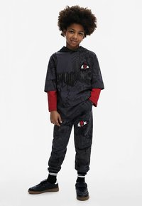 Musta ja harmaa hupullinen asu salaman kuvioilla, alla punaiset pitkät hihat, ja paidassa sekä housuissa graafiset kangasmerkit. Mukana mustat lenkkarit.