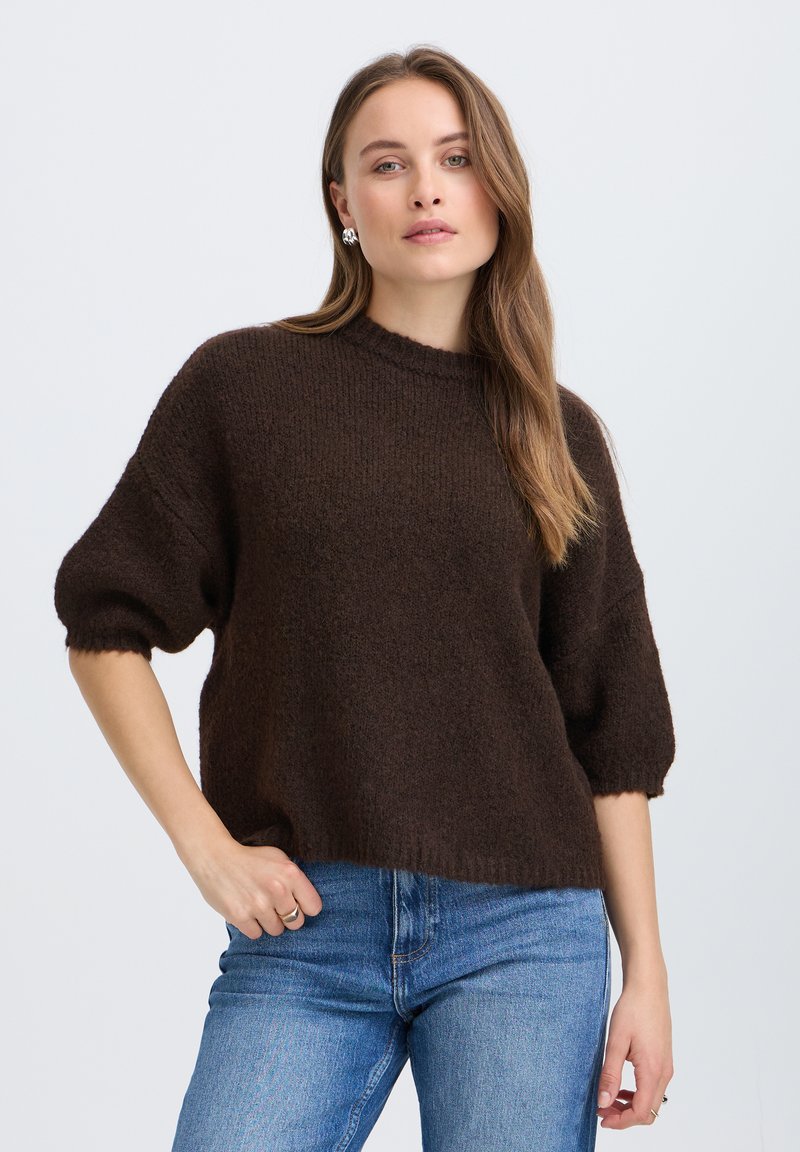 Femme aux longs cheveux bruns portant un pull texturé marron foncé avec des manches aux coudes et un jean bleu sur un fond uni.