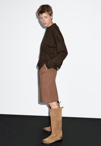 Maglione lavorato a maglia marrone, pantaloni beige e stivali al ginocchio in suede color nocciola con dettagli a anello, su uno sfondo bianco.