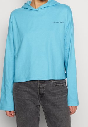 Osoba nosząca jasnoniebieską krótko skrojoną bluzę z kapturem z napisem "MARCO'POLODENIM" na piersi oraz wyblakłe czarne dżinsy.