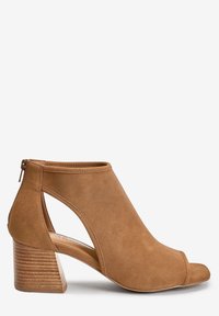 Bottines ouvertes en daim beige, avec design à découpes sur le côté, fermeture éclair et talon en bois empilé. Texture lisse sur l'ensemble.