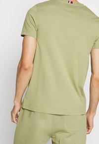 Camiseta verde oliva con escote redondo y mangas cortas, que presenta un pequeño detalle en rojo, blanco y azul en la parte posterior del cuello.