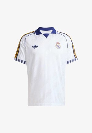 Bela nogometna majica z modrim ovratnikom, rumenimi in modrimi črtami na rokavih, logotipom Adidas na levi strani prsnega koša in grbom Real Madrida na desni strani prsnega koša.