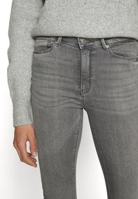 Jeans de denim cinza com um corte justo, apresentando um design clássico de cinco bolsos, leve desbotamento e um botão prateado na cintura.