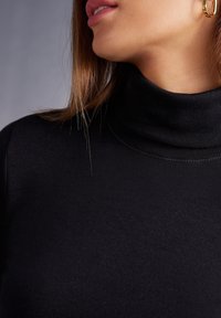 Pull à col roulé noir en tissu tricoté lisse, avec un col ajusté et des détails de couture subtils. Boucle d'oreille dorée visible.
