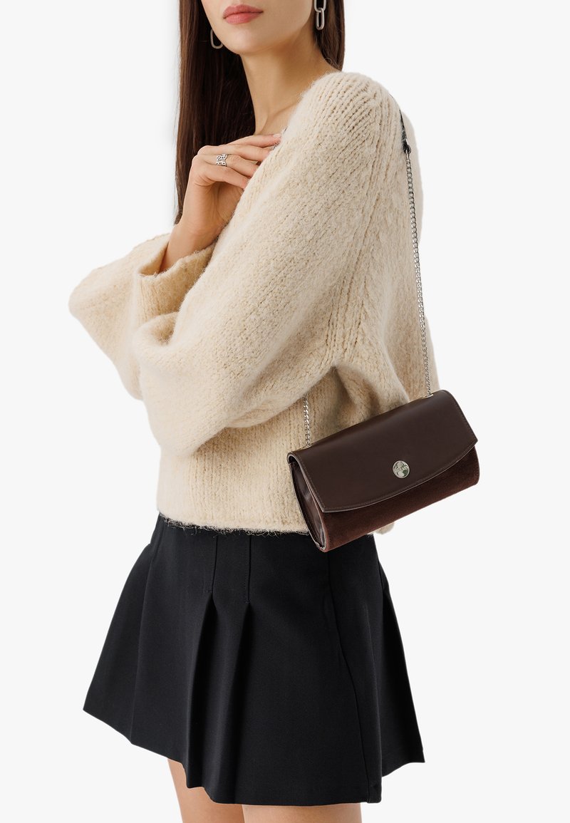 Brązowa skórzana torebka na ramię z zamszu, z srebrnym łańcuchem, posiadająca zaokrągloną klapkę i okrąły akcent z logo.