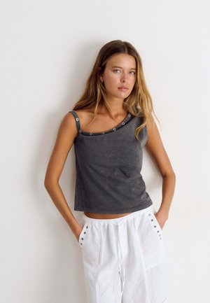 Mujer joven con cabello largo y rubio lleva una camiseta sin mangas gris con ojales y pantalones blancos con manos en los bolsillos contra un fondo liso.