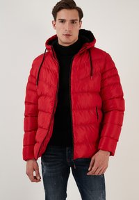 Rød polstret jakke med hætte, lynlås lukning og sorte detaljer. Har quiltet design og forlommer, båret over en sort sweater.