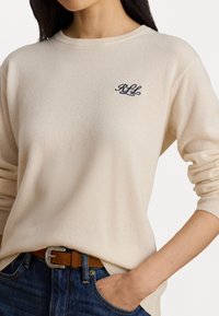 Pullover de malha creme com decote redondo e punhos canelados, apresentando um logótipo bordado em azul escuro no peito. Usado com jeans de ganga.