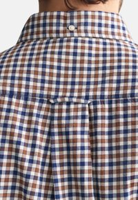 Camisa a cuadros en tela marrón, azul y blanca; presenta un collar clásico, detalle de botones y un pliegue en la parte trasera para mayor libertad de movimiento.