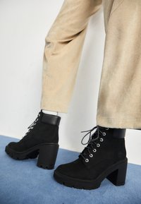 Black suede ankle boots with a chunky heel, round toe, and silver eyelets, featuring a matte black leather collar and textured sole.