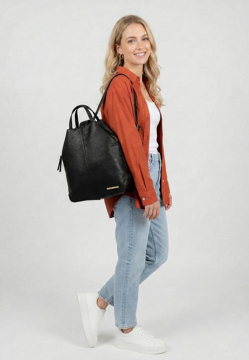 Femme souriante portant une chemise orange, un haut blanc, un jean bleu clair, des baskets blanches, portant un grand sac à dos en cuir noir sur l'épaule devant un fond gris.