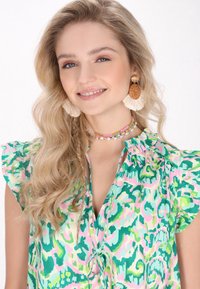 Színes zöld és rózsaszín mintás blúz puffos ujjal és megkötős elülső résszel. Párhuzamos, kör alakú fülbevalók és gyöngyös choker ékszer díszíti.