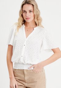 White short-sleeve blouse with eyelet pattern, V-neck, button front, and smocked waist, paired with tan pants. Smooth texture.