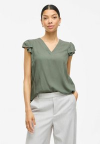 Blusa verde oliva con escote en V y mangas cortas con acentos fruncidos. Textura suave, corte holgado, combinada con pantalones gris claro.