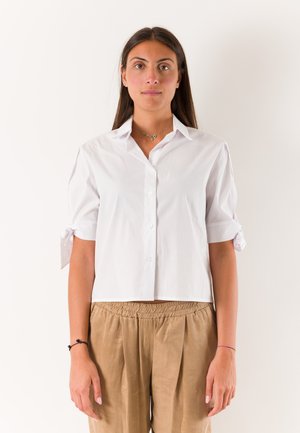 Giovane donna con capelli lisci e scuri che indossa una camicia bianca a maniche corte abbottonata e pantaloni larghi marrone chiaro, in piedi davanti a uno sfondo bianco.