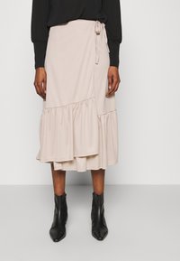Femme portant une jupe midi beige à enrouler avec des détails de niveau froncé et des bottines noires à bout pointu, se tenant contre un fond neutre.