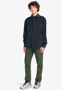 Camicia in corduroy blu navy con chiusura a bottoni, colletto e taschino sul petto, abbinata a pantaloni verdi e scarpe da ginnastica argento.