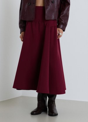 Donna che indossa una gonna midi bordeaux, giacca di pelle scura, stivali neri e un anello d'argento, in piedi davanti a uno sfondo chiaro.