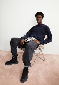 Marineblauw sweatshirt met een klein logo, relaxed-fit donkergrijze jeans en zwarte high-top laarzen, zittend op een zilveren opklapstoel tegen een lichte achtergrond.