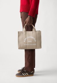 Beige lærred tote bag med hvid tekst, der har to robuste håndtag, rektangulær form og et minimalistisk design. Båret af en person i brune bukser.