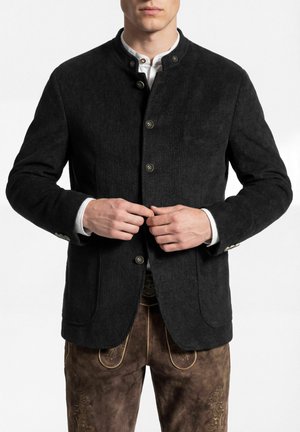 Mann knöpft schwarze Cordjacke über weißem Hemd, trägt bestickte braune Lederhose, vor schlicht weißem Hintergrund.