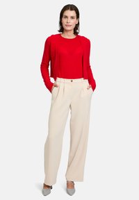 Cardigan ajusté rouge sur un haut rouge, accompagné d'un pantalon beige taille haute et large avec des plis sur le devant et une fermeture à un bouton.