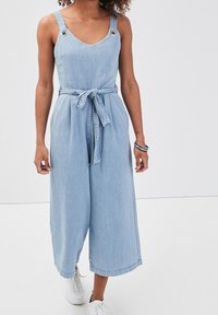 Light blue denim jumpsuit with wide legs, V-neckline, adjustable shoulder straps, and a fabric belt. Features silver-toned hardware accents.
