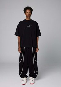 Zwarte oversized t-shirt met wit scriptlogo, gecombineerd met zwarte broek met witte piping. Witte sneakers met rode accenten.