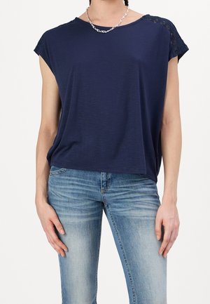Granatowy top z krótkim rękawem o miękkiej fakturze z koronkowymi akcentami na ramionach, w połączeniu z dopasowanymi jasnoniebieskimi jeansami.