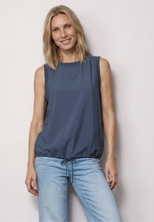 Femme aux cheveux blonds mi-longs portant un haut bleu sans manches et un jean bleu clair, souriante et debout devant un fond uni.
