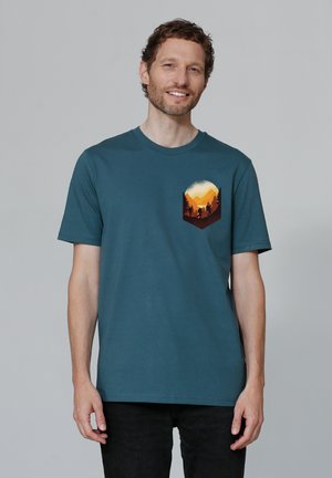 Teal katoenen t-shirt met een ronde hals. Beschikt over een hexagonaal zakprint van een zonsondergang boven bergen en bomen in warme kleuren.