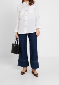 Camisa blanca semitranslúcida de botones con dos bolsillos en el pecho, combinada con pantalones vaqueros oscuros de pierna ancha y bailarinas de estampado de leopardo. Bolso negro.