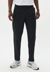 Pantaloni neri sartoriali con una texture liscia e pieghe frontali, abbinati a scarpe sportive grigie e blu con tomaia in rete e suola imbottita.