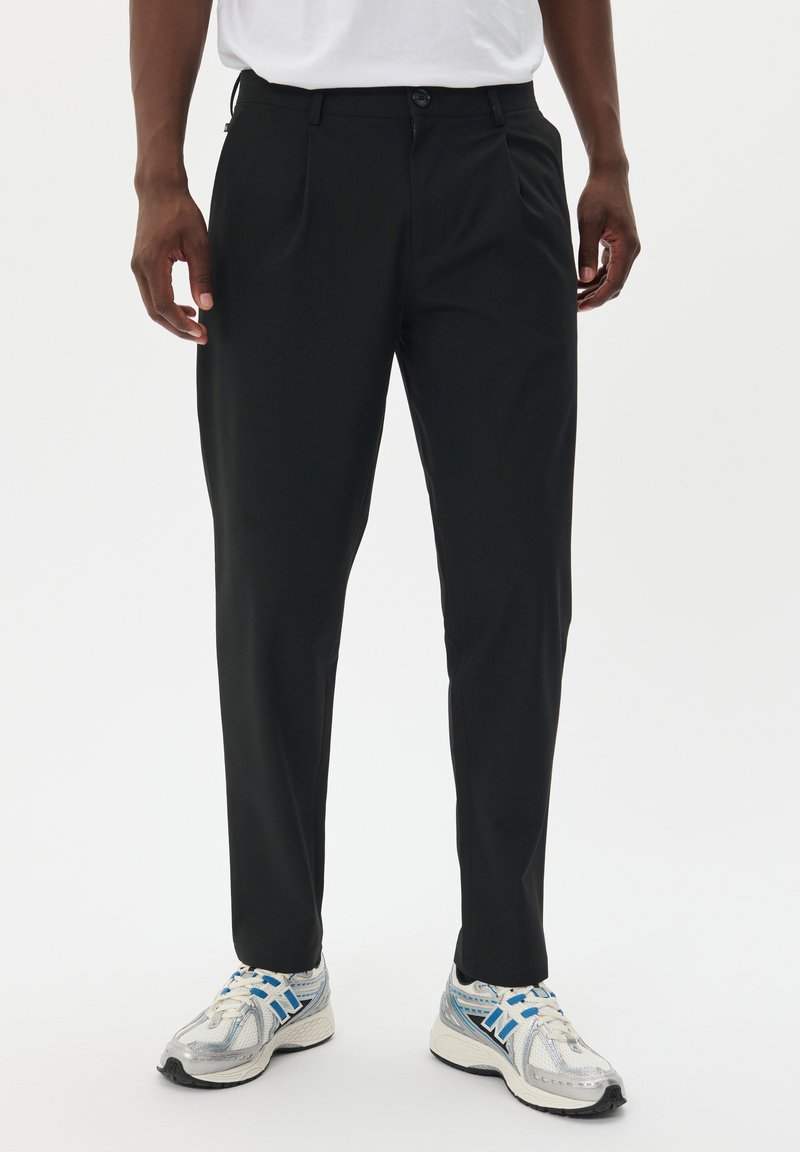 Pantaloni neri sartoriali con una texture liscia e pieghe frontali, abbinati a scarpe sportive grigie e blu con tomaia in rete e suola imbottita.