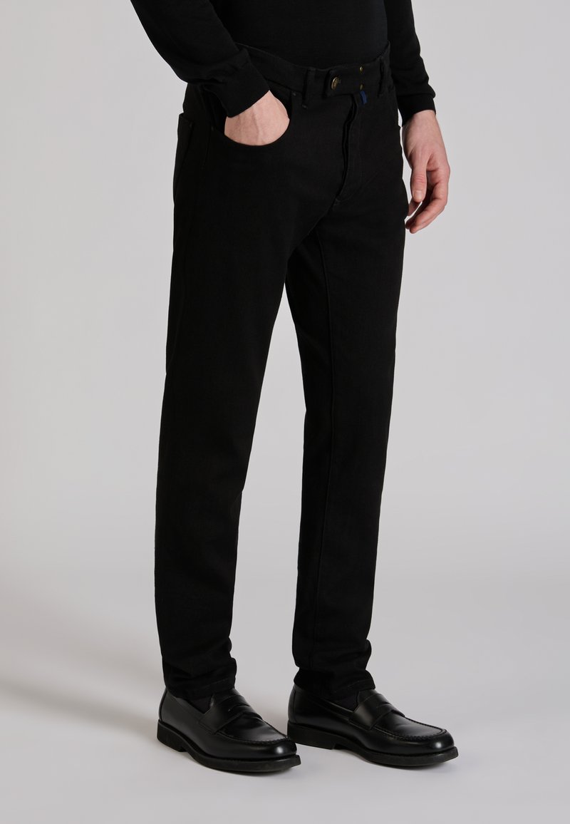 Man die zwarte slim-fit broek, zwart shirt met lange mouwen en zwarte nette schoenen draagt, staat met één hand in de zak.