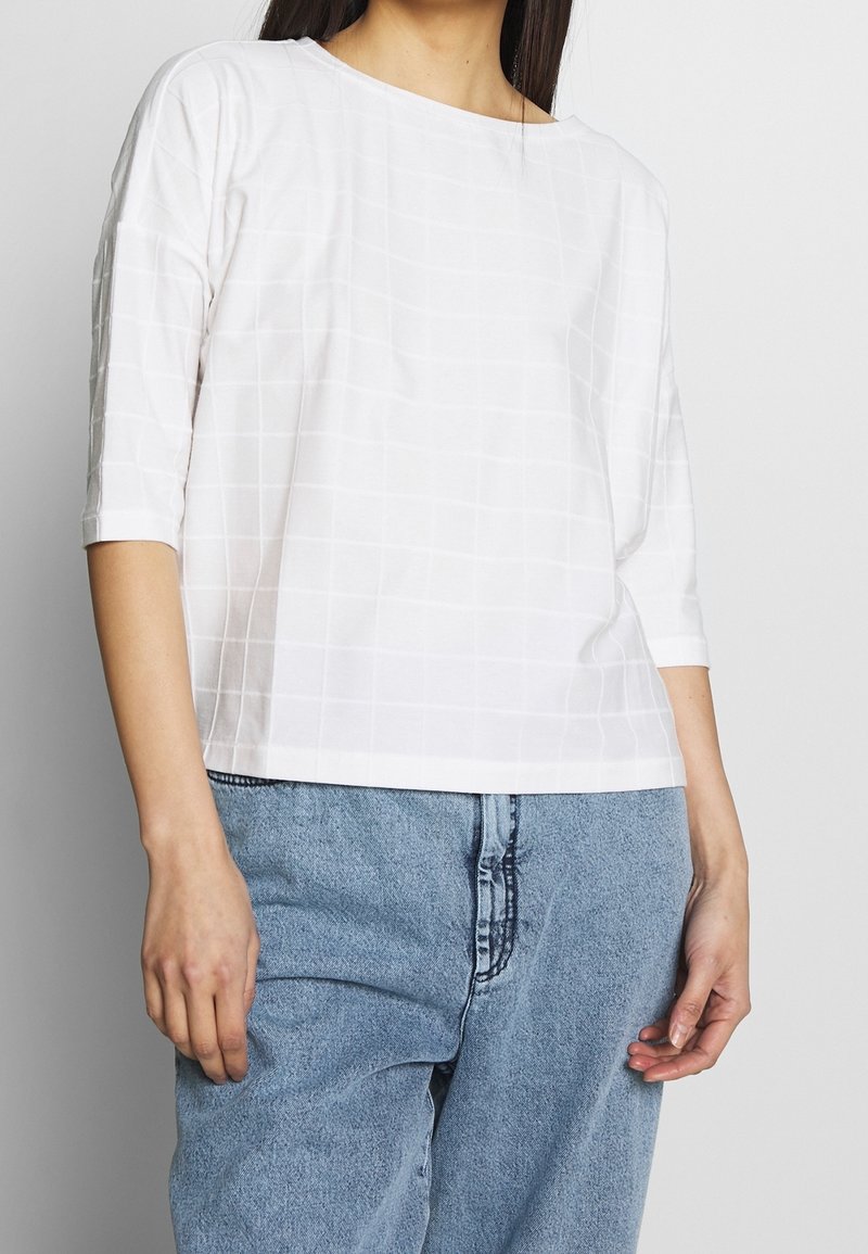 Femme portant un haut blanc à manches trois-quarts avec un motif quadrillé subtil et un jean taille haute bleu clair sur un fond uni.