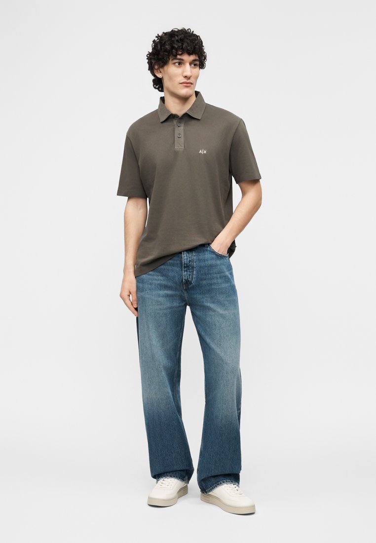 Homme portant un polo olive foncé, un jean bleu ample et des baskets blanches, debout avec une main dans la poche sur un fond uni.