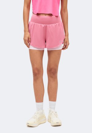 Kvinde iført pink lagdelte atletikshorts, hvide sokker og hvide løbesko, stående mod en ensfarvet baggrund.