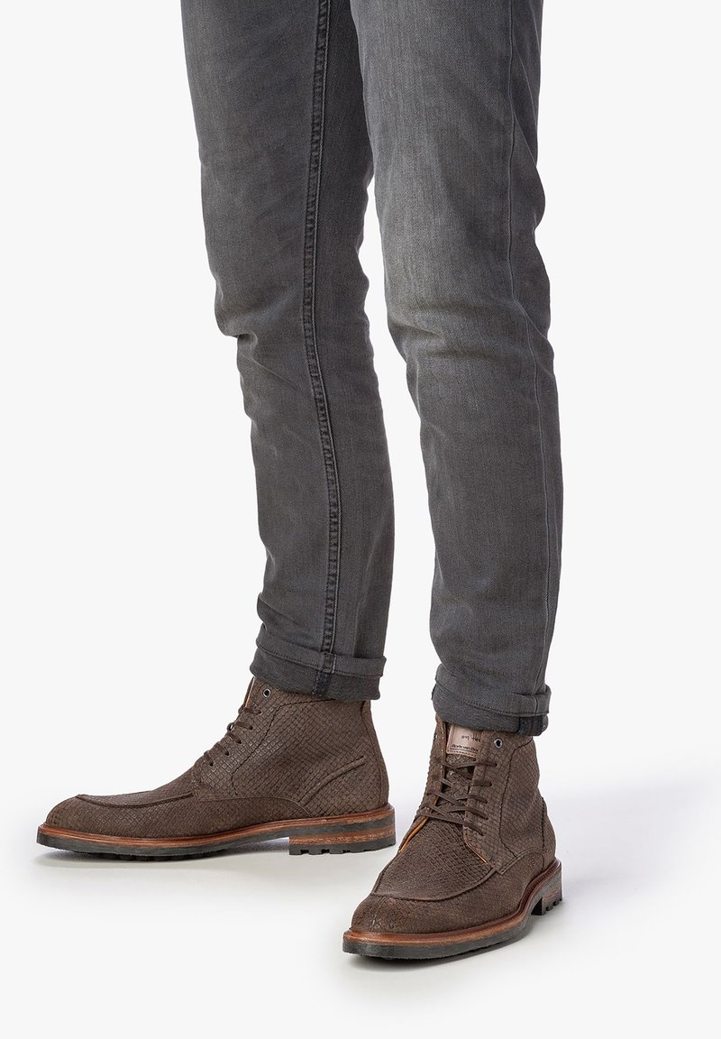 Botas marrones con textura, con cordones y una superficie estampada, acompañadas de una suela de goma en contraste, combinadas con pantalones oscuros de mezclilla ajustados.