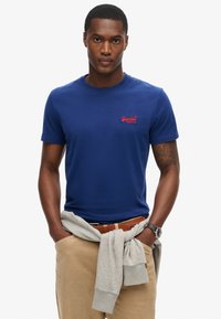 Camiseta de algodón azul marino con cuello redondo, que presenta un pequeño logo rojo en el pecho. Pantalones beige con un cinturón marrón, sudadera gris atada a la cintura.