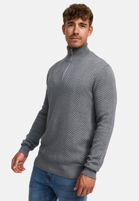 Grå strikket genser med høy glidelås og krage, med en teksturert herringbone-mønster. Ribbestrikkede mansjetter og nederkant gir en tett passform.