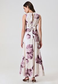 Robe maxi en tissu crème avec un imprimé floral violet. Design sans manches, détail en trou de serrure au dos et ceinture à la taille. Chaussures à talons visibles.
