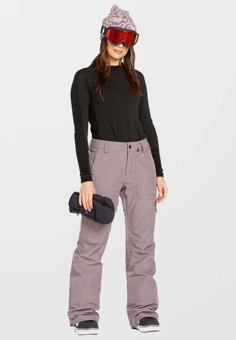 Conjunto de esquí que incluye una camiseta ajustada de manga larga negra, pantalones de esquí morados claros con bolsillos y botas de nieve negras con detalles en plata; sosteniendo guantes.