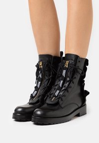 Bottes en cuir noir montantes, avec fermetures éclair dorées, lacets en ruban et semelle robuste. Coutures détaillées sur les côtés et design à lacets.