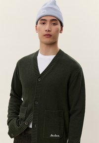 Νεαρός άνδρας που φοράει γαλάζιο σκουφάκι, λευκή μπλούζα και σκούρο πράσινο πουλόβερ με κουμπιά και μπροστινές τσέπες, στέκεται μπροστά σε μπεζ φόντο.