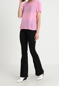 Różowa bluzka z krótkim rękawem z miękkiego, przezroczystego materiału, w połączeniu z czarnymi spodniami dzwonowymi i białymi, szpiczastymi botkami za kostkę.