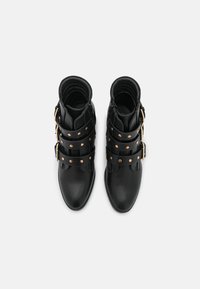 Pár černých kožených kotníkových bot s třemi širokými pásky a zlatými sponami, viděno zhora na bílém pozadí.