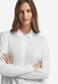 Vit stickad knapp-up cardigan, med krage, långa ärmar och ett texturerat mönster genom hela plagget. Fyra knappar synliga på placket.