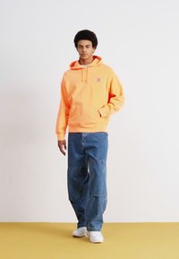 Oranje hoodie met een zak en logo-patch, gecombineerd met losse, lichtblauwe denim broek. Het model draagt witte sneakers. Effen achtergrond.